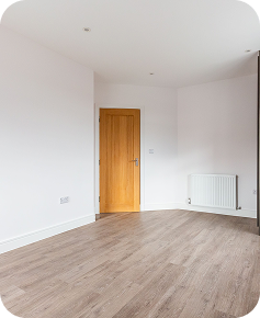 A bright, empty room with white walls, a wooden floor, a closed light wood door, a white radiator, and minimal furnishings. Natural light brightens the clean, modern space.