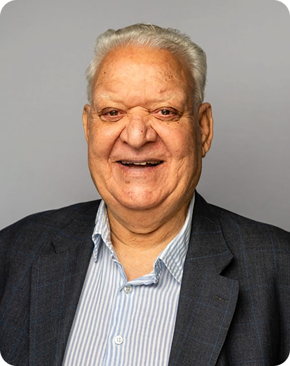 An elderly man with short white hair smiles warmly, wearing a navy blazer over a light blue striped shirt, posing against a plain gray background.