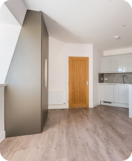 A modern, unfurnished kitchen with light wood flooring, white cabinets, a stainless steel sink, and a wooden door. Natural light brightens the space, and a dark feature wall stands beside the door.