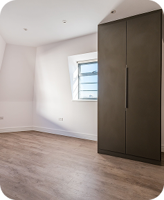 A bright, empty room with light wood flooring, white walls, a large dark wardrobe, and a single window letting in natural light.