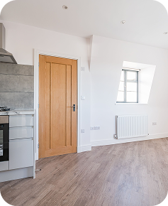 A modern, bright kitchen with wooden flooring, a light wood door, a small window, white walls, a radiator, and part of a stove and countertop visible on the left.