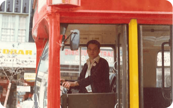A man wearing a dark jacket is sitting in the drivers seat of a red double-decker bus, looking out the window. The bus is parked on a city street with shop signs visible in the background.
