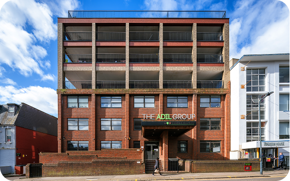 A modern, multi-story building with a brick and concrete facade, large balconies, and The Adil Group sign above the entrance. The sky is blue with scattered clouds.