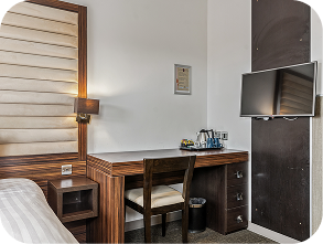Modern hotel room corner with a wooden desk, chair, wall-mounted TV, tea set, lamp, and part of a bed visible on the left. The space appears neat and well-lit.