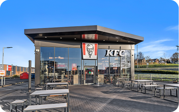 A modern KFC restaurant with large glass windows, outdoor seating, and the KFC logo featuring Colonel Sanders above the entrance, set on a clear, sunny day.