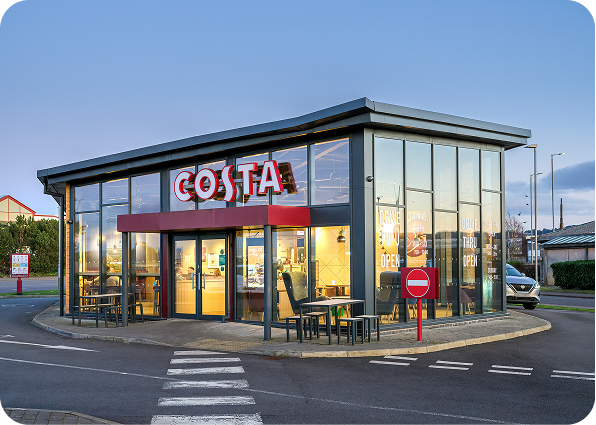 A modern Costa Coffee shop with large glass windows, red signage, and outdoor seating is shown at dusk. The building is surrounded by a drive-through lane with a no entry sign visible in the foreground.
