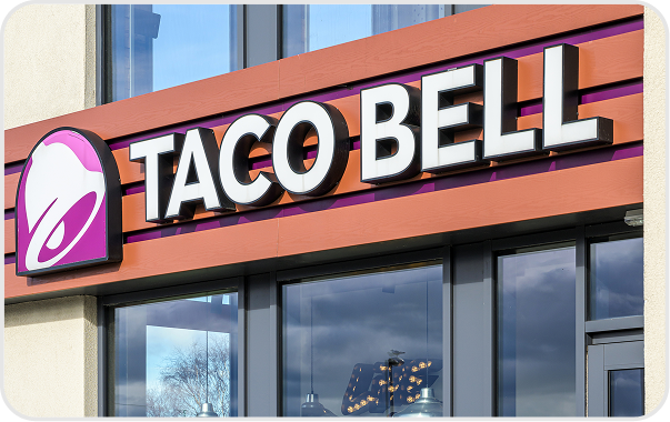 The image shows the exterior sign of a Taco Bell restaurant, featuring the Taco Bell logo and bold white letters on a brown and orange background above large windows.