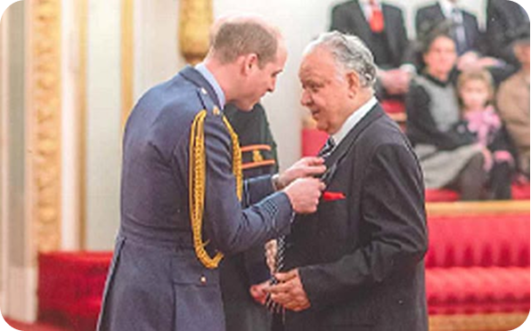A man in a military uniform presents an award or medal to an older man in a suit inside an ornate room, while people seated in the background observe the ceremony.