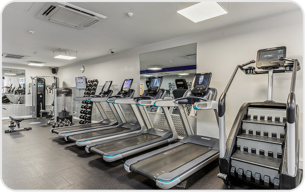 A modern gym with treadmills, a stair machine, free weights, an exercise bike, and various strength training equipment. The space is clean, well-lit, and has mirrors along one wall.