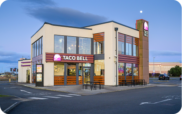A modern Taco Bell restaurant with large windows, outdoor seating, and illuminated Taco Bell signs at dusk, under a clear sky with the moon visible.