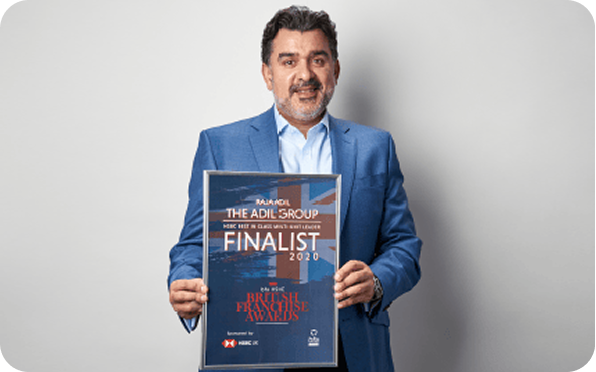 A man in a blue suit smiles while holding a framed certificate that reads The Adil Group Finalist 2020, British Franchise Awards. He stands against a plain light background.