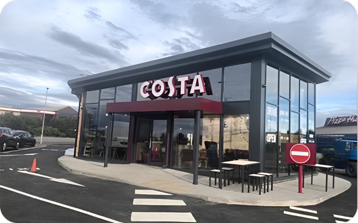A modern glass-fronted Costa coffee shop with outdoor seating. A red awning and large COSTA sign are above the entrance. A no entry sign is by the sidewalk, and parked cars are visible nearby under a cloudy sky.