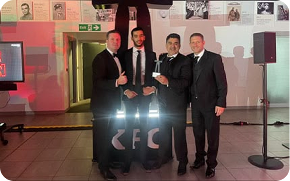Four men in tuxedos stand together indoors at an event, smiling and holding an award. Behind them is a large KFC logo and display with photos and text on the wall.