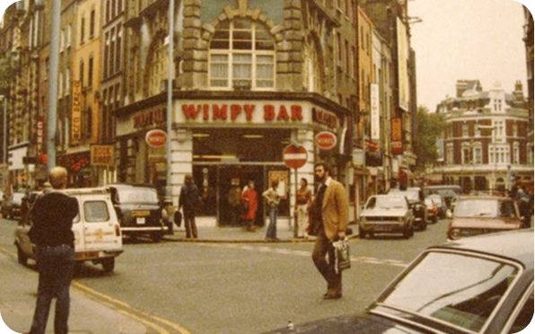 A busy city street scene from the 1970s with people crossing the road, vintage cars parked, and a Wimpy Bar restaurant on a corner building surrounded by tall, older-style architecture.