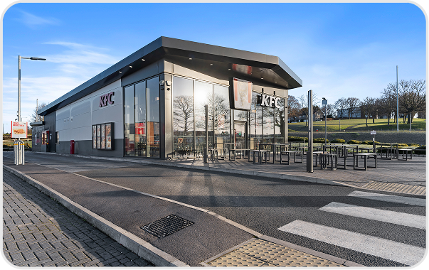 A modern KFC restaurant with large glass windows and outdoor seating is shown on a sunny day. The area is clean and empty, with a crosswalk and paved road in front. Trees and open green space are visible in the background.