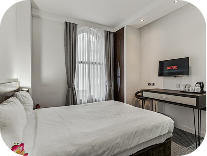 A bright, modern hotel room with a large bed, white linens, a window with gray curtains, a wall-mounted TV, and a desk with a kettle and cups. The room features minimalistic decor and neutral colors.