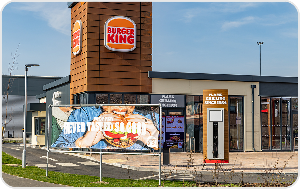 A Burger King restaurant with a prominent logo on a tall sign and a banner outside showing a person holding a large burger with the text NEVER TASTED SO GOOD. The building has modern architecture and clear skies above.