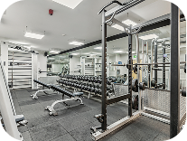 A modern gym with weight racks, dumbbells, benches, and squat racks. The room has large mirrors on the walls, bright lighting, and a clean, organized appearance.