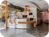 A modern hotel reception area with a curved wooden front desk, two computer monitors, decorative lighting, a staircase, and a patterned accent wall in the background.