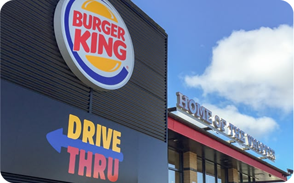 The image shows a Burger King restaurant with a large logo and a Drive Thru sign. The building is under a blue sky with some clouds, and part of the Home of the Whopper slogan is visible.