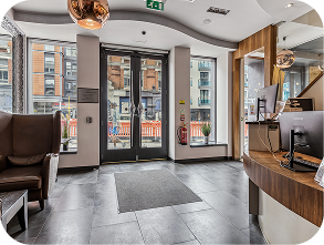 Modern hotel lobby with large glass doors, reception desk, seating area, and city buildings visible outside through the windows.