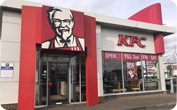 Exterior of a KFC restaurant with a large Colonel Sanders logo, glass doors, and a sign indicating it is open till 2 AM on Fridays and Saturdays. Some tables and chairs are visible through the windows.