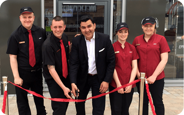 A group of five people, including a man in a suit cutting a red ribbon, stand in front of a building. Four others, wearing matching work uniforms, smile beside him, marking an opening ceremony.