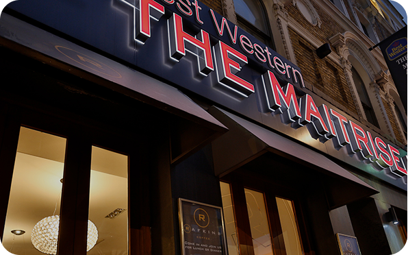 A nighttime view of the illuminated sign for Best Western THE MAITRISE hotel, with warm lights glowing from the windows and part of a restaurant sign visible below.
