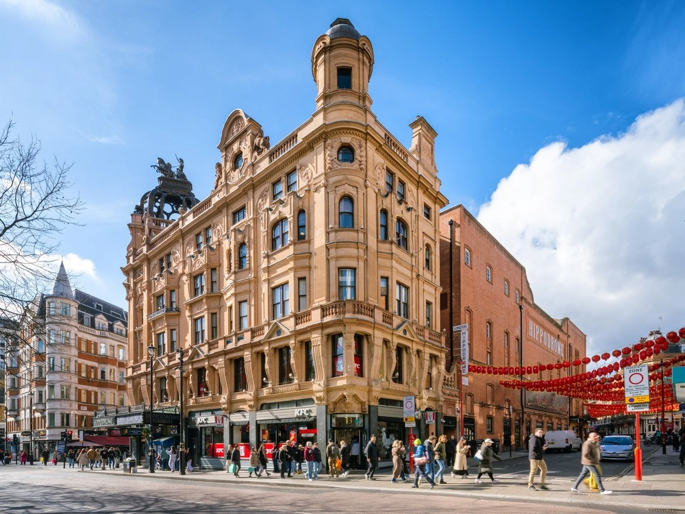 A busy London street scene with people walking past an ornate, historic beige building under a blue sky; red lanterns hang on the right, marking the entrance to Chinatown.