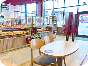 A bright, modern bakery café with large windows, a display counter filled with pastries and snacks, wooden tables and chairs, and a clear view of the parking lot outside.