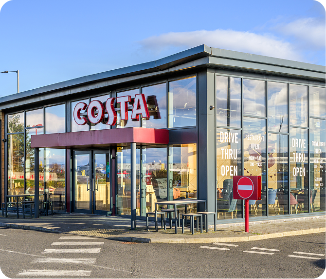 Modern Costa Coffee shop with large glass windows, outdoor seating, and a drive-thru. A red and white Costa sign is above the entrance, and a no entry traffic sign is visible in the foreground.