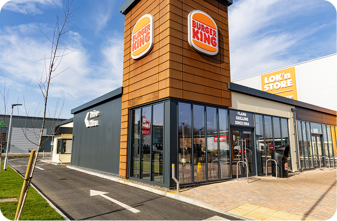 A modern Burger King restaurant with large glass windows and a drive-thru lane is shown on a bright day. The building features the Burger King logo and is next to a Lok’nStore facility.