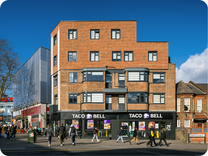 A Taco Bell restaurant occupies the ground floor of a modern brick building on a busy street; several people walk past on the sidewalk under a blue sky.