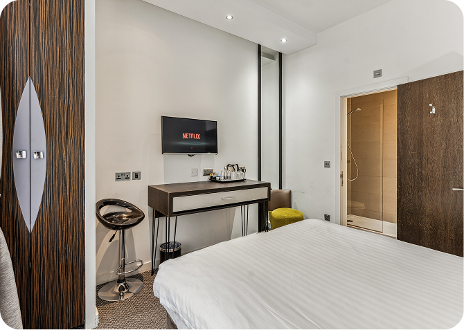 Modern hotel room with a bed in the foreground, a wall-mounted TV displaying the Netflix logo, a desk with a chair and tea set, a tall wardrobe, and an open door showing a bathroom with a shower.
