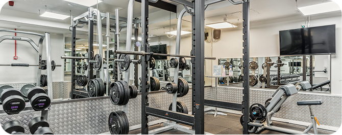 A gym room with weightlifting equipment, including dumbbells, barbells with weight plates, squat racks, benches, mirrors on the walls, and a mounted flat-screen TV.