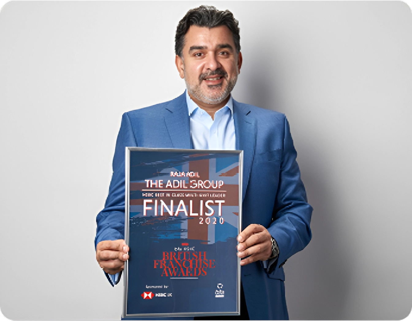 A man in a blue suit smiles while holding a framed certificate for being a 2020 finalist at the British Franchise Awards, representing The Adil Group.