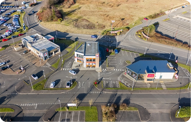 Aerial view of a commercial area with three fast-food restaurants, parking lots, and several cars. Surrounding roads curve around the buildings, and there is an open grassy field in the background.