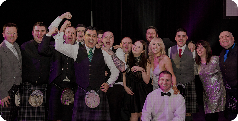 A group of people dressed in formal attire, some wearing kilts, stand together smiling and posing enthusiastically for a group photo at an event or celebration.