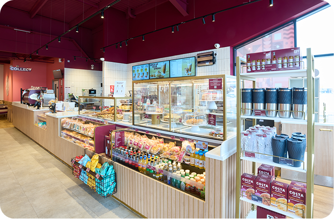 A modern café with a maroon and white interior features glass display cases filled with pastries, sandwiches, and drinks. Shelves hold coffee cups and merchandise, and a barista works behind the counter.