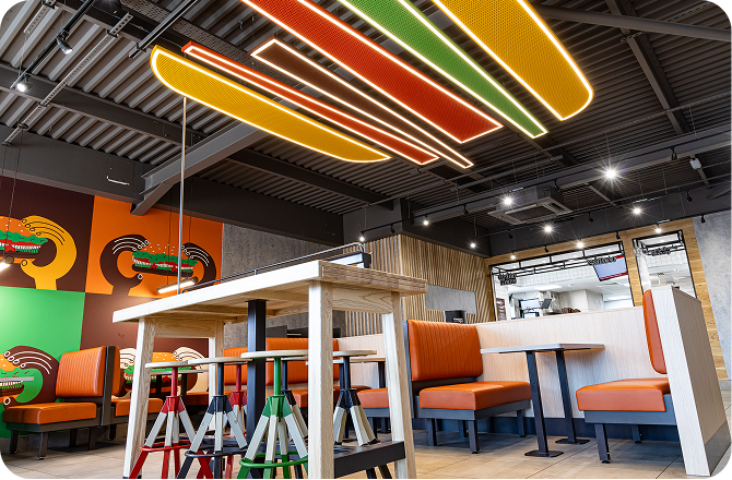 A modern fast-food restaurant interior with orange booths, a wooden table, colorful stools, and bright neon ceiling lights; bold wall art features abstract designs in green, orange, and yellow.