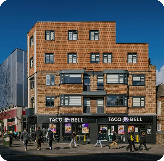 A red-brick building with a Taco Bell restaurant on the ground floor. Several people are walking on the sidewalk in front, and the sky is clear and blue.