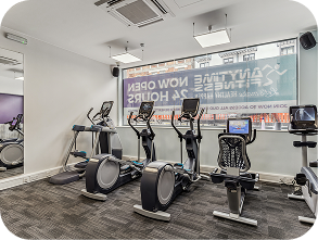 Small gym room with three elliptical machines and one recumbent exercise bike facing a large window. The window displays a sign for Anytime Fitness and Now Open 24 Hours. The room has grey carpet and white walls.
