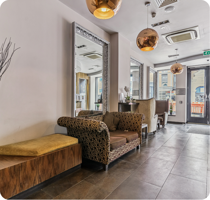 Modern waiting area with a patterned couch, a cushioned wooden bench, large mirrors, stylish pendant lights, and two armchairs by tall windows. The space has a tiled floor and neutral-toned decor.