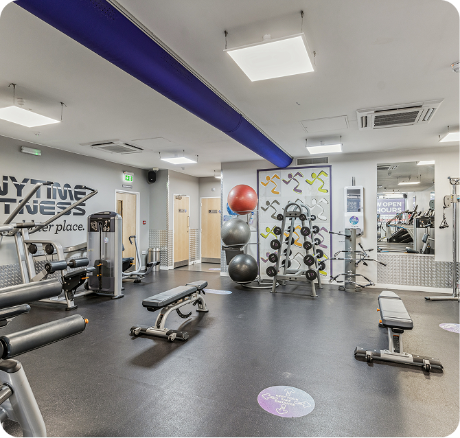 Modern gym with weight benches, exercise machines, a rack of colorful medicine balls, wall-mounted weights, and mirrors. The floor has social distancing markers and the walls display gym branding and motivational quotes.