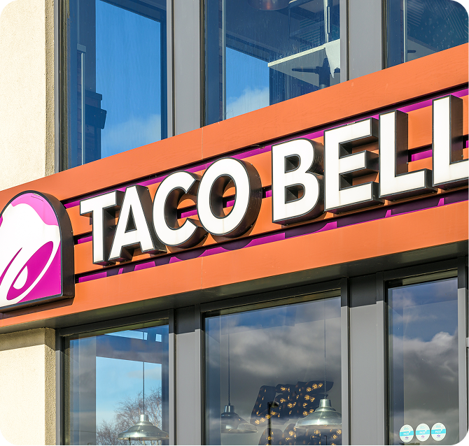 The exterior of a Taco Bell restaurant featuring its logo, large sign with bold white letters, and glass windows reflecting the sky and clouds.