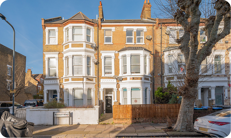 A three-story Victorian-style brick townhouse with white trim and bay windows, located on a sunny residential street with a street sign reading “Buckley Road” and leafless trees in front.