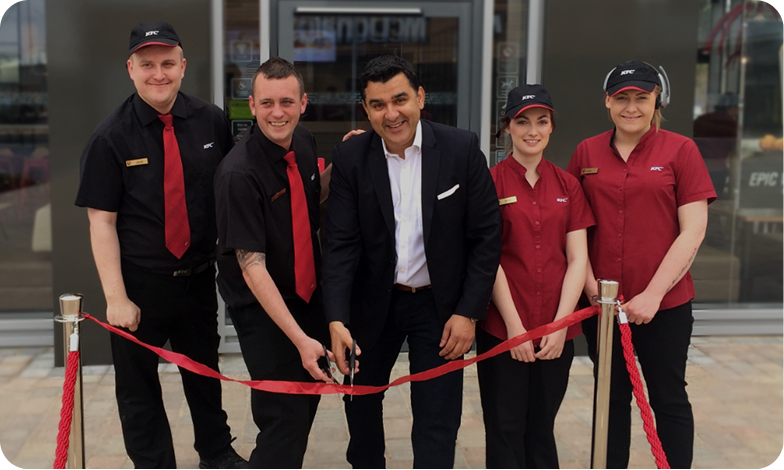 Five people, four in KFC uniforms and one in a suit, stand in front of a building smiling. The person in the suit is cutting a red ribbon, suggesting a store opening or special event.