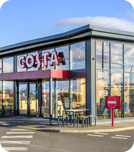 A modern Costa Coffee shop with large glass windows and a red sign, featuring outdoor seating and a Drive Thru Open sign. A no entry traffic sign is visible in the foreground.