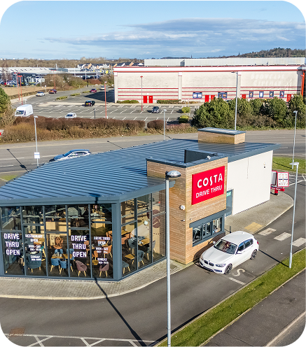 A white car at the drive-thru window of a modern Costa Coffee building with large glass windows. A red COSTA DRIVE THRU sign is visible, and the drive-thru is open. Surrounding roads and an industrial area are in the background.