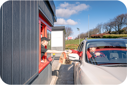 A person in a car receives food from an employee at a fast-food drive-thru window on a sunny day. The employee hands over a KFC bag as the driver reaches out through the car window.